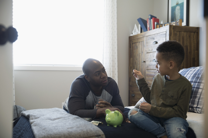 Father and son depositing coins into piggy bank in bedroom