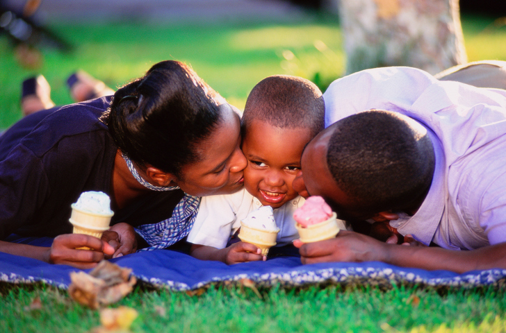 Family lying on grass with ice-creams, parents kissing boy's face