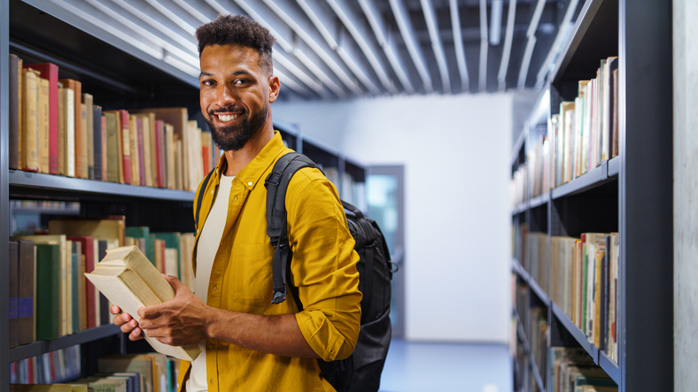 Young university student with book indoors in librabry, looking at camera.