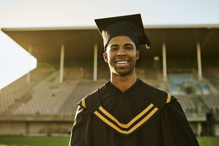 Portrait of smiling graduate in gown and hat standing alone on university campus at graduation ceremony. Excited, happy qualified postgrad graduating college for academic bachelor, masters and degree