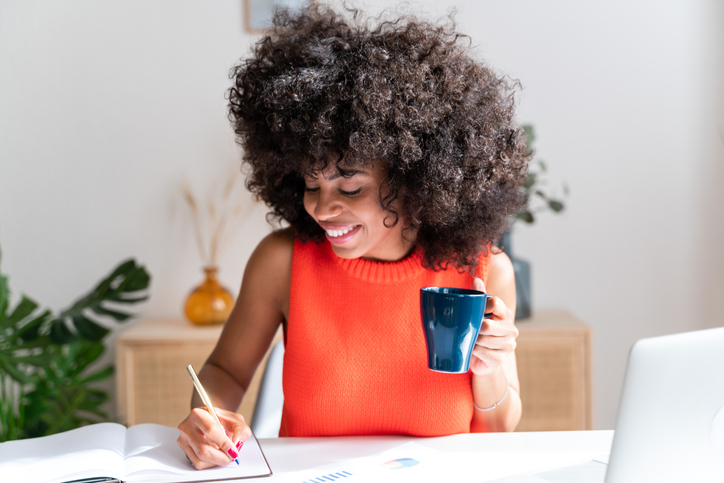 smiling african american woman working from home smiles as she holds a cup of coffee and takes some notes in a notebook
