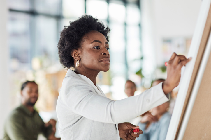 Creative black woman, meeting and writing on whiteboard in planning, brainstorming strategy or coaching at office. African American female in presentation using board to write idea or FAQ for startup