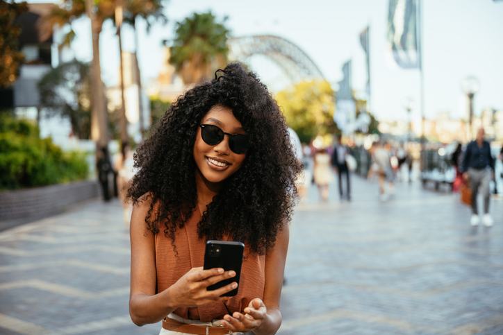Smiling woman with curly hair and sunglasses using smart phone in sunny urban park with people walking.