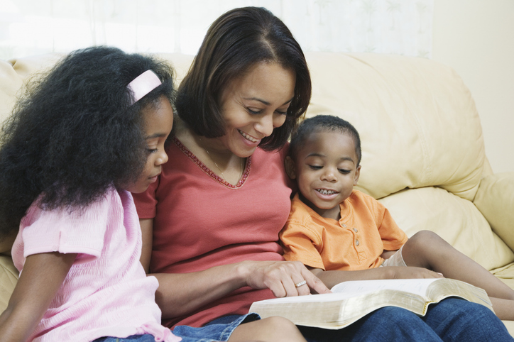 Mother with children (4-7) on sofa, reading bible, smiling, Mother's Day