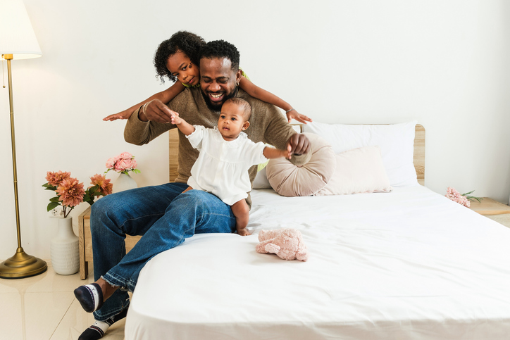 A joyful family plays together on a bed, showcasing love and connection in a bright, cozy room with soft decor. A joyful father enjoys playful moments with his baby and young daughter on a cozy bed.