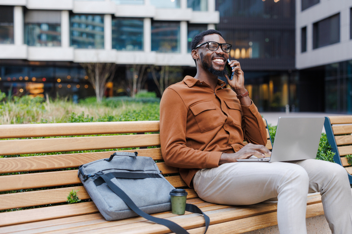 Smiling man working remotely on a laptop while talking on the phone outdoors