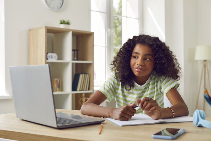 Happy school girl sitting at her desk at home, having online class and looking at laptop