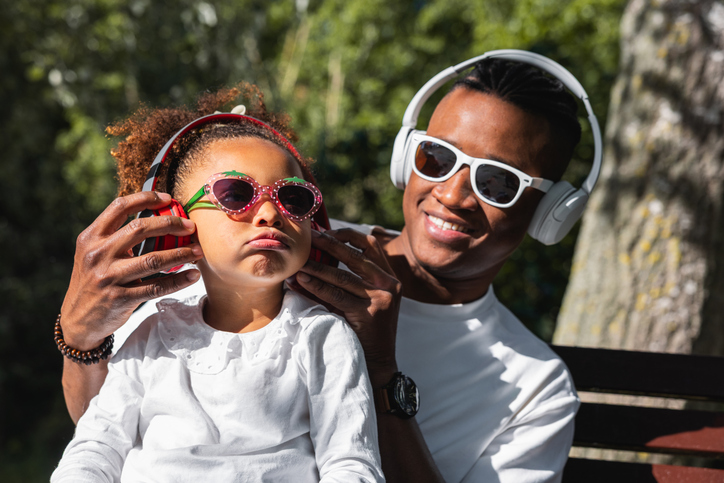 Father and daughter listening to music outdoors in the park, both wearing headphones and sunglasses. The girl seems to dislike the music her father has chosen.