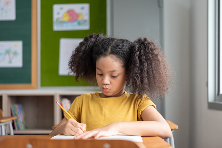 African American girl with curly hair writes in her notebook at her school desk during a lesson.
