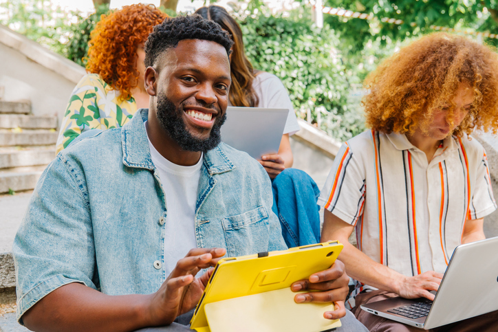 Diverse college students lounging on campus steps, engaging with digital devices, sharing laughter and camaraderie during study break