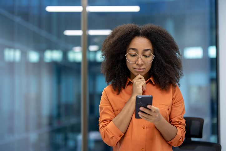 Serious thinking woman standing with phone in hands near window inside office. Businesswoman reading online news and browsing internet pages at workplace, using app