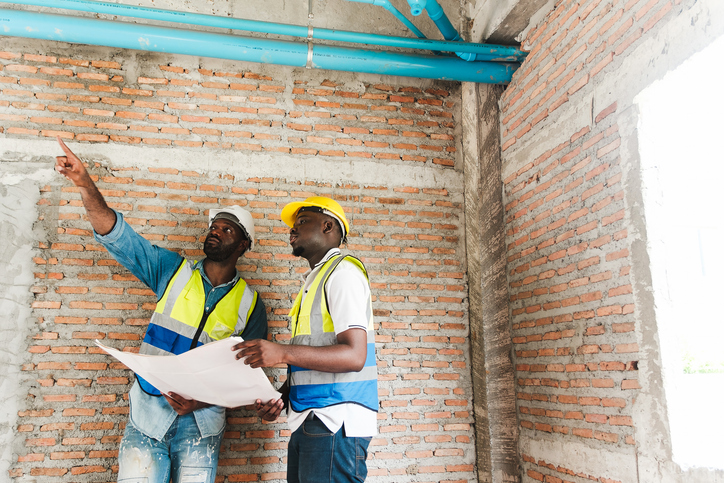 Two construction workers in safety gear inspect a brick wall with exposed piping on-site, reviewing blueprints and discussing installation, planning progress for a building or housing project.