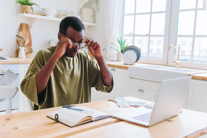 Young man is adjusting his glasses and reviewing his finances at the kitchen table, with a laptop and calculator in front of him