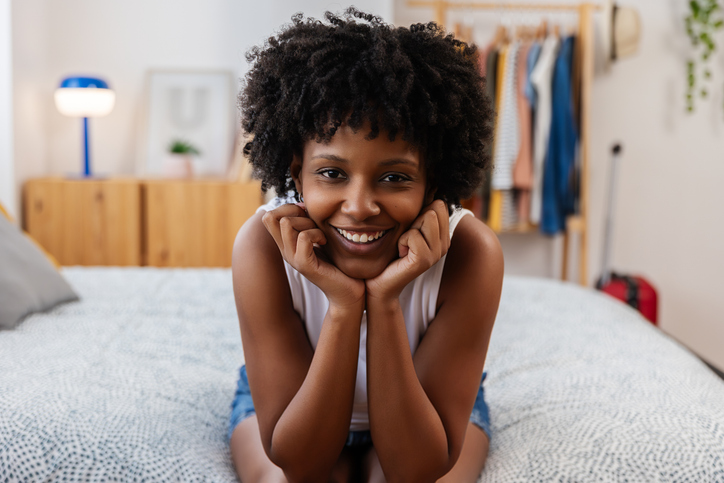 Portrait of young black woman smiling at camera and relaxing on bed in bedroom