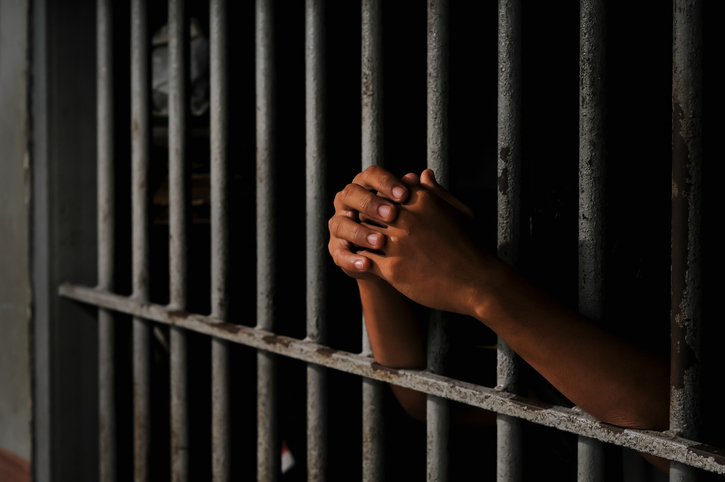 Close-up of an inmate’s hand holding prison bars in a dark cell.