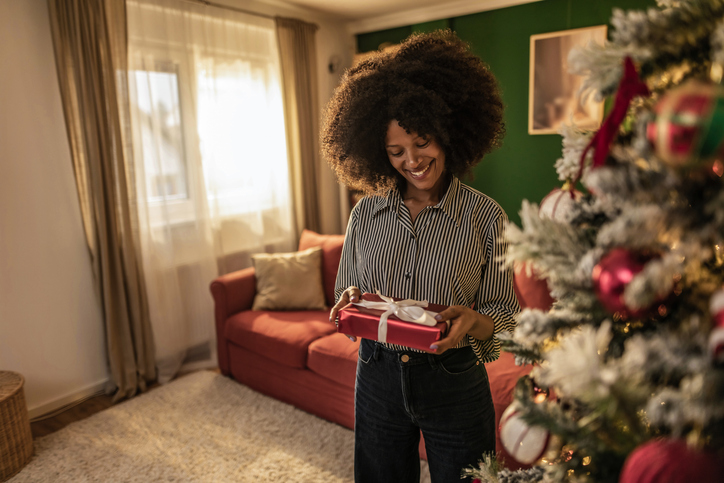Young black woman holding a Christmas present next to Christmas tree at home